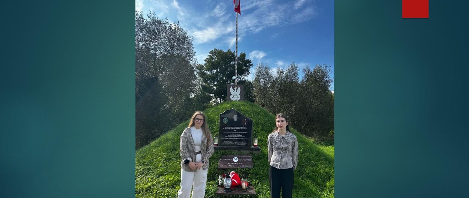 Dwie dziewczyny stoją przy pomniku, przy którym złożona jest wiązanka kwiatów oraz znicze. W tle kopiec, a na jego szczycie maszt z biało-czerwoną flagą.
