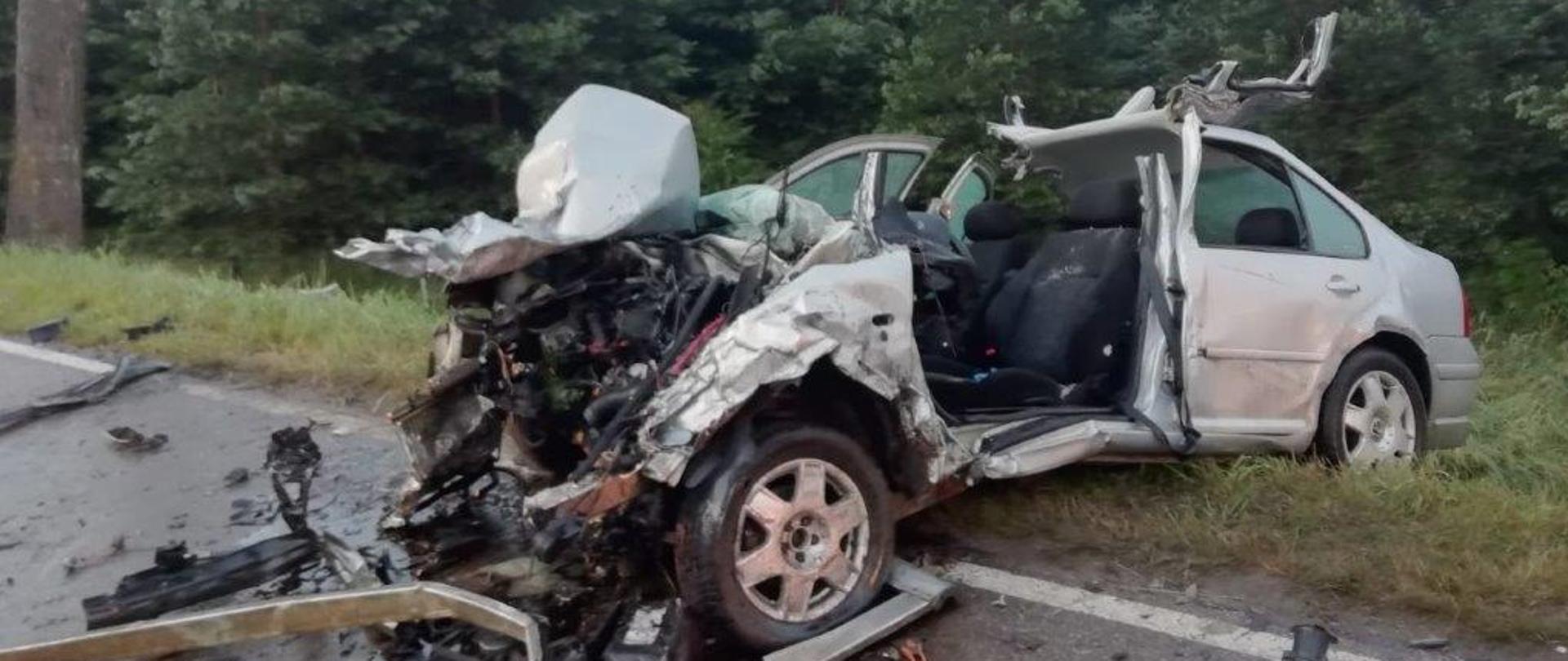 Zdjęcie przedstawia rozbity samochód osobowy znajdujący się częściowo poza pasem jezdni