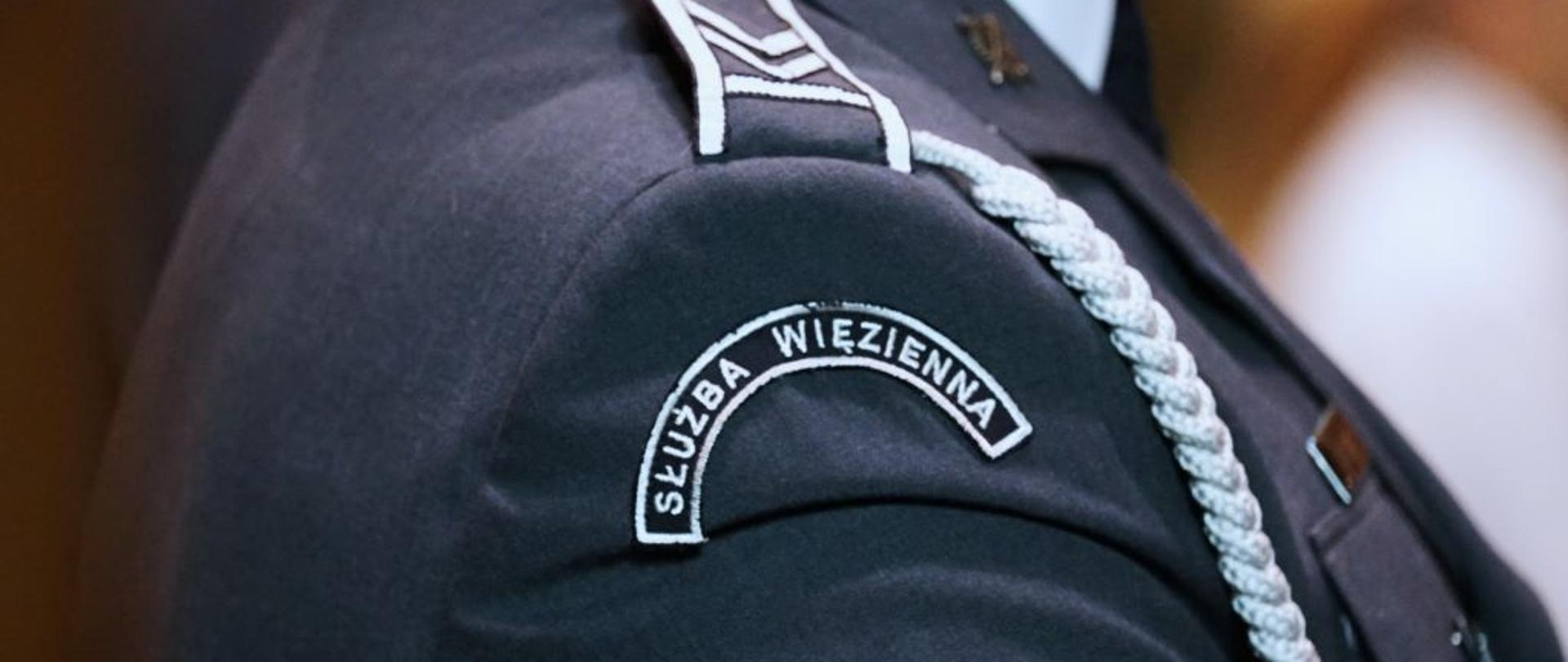 ramię człowieka w srebrnym mundurze . na ramieniu napis służba więzienna oraz pagon ze stopniem 