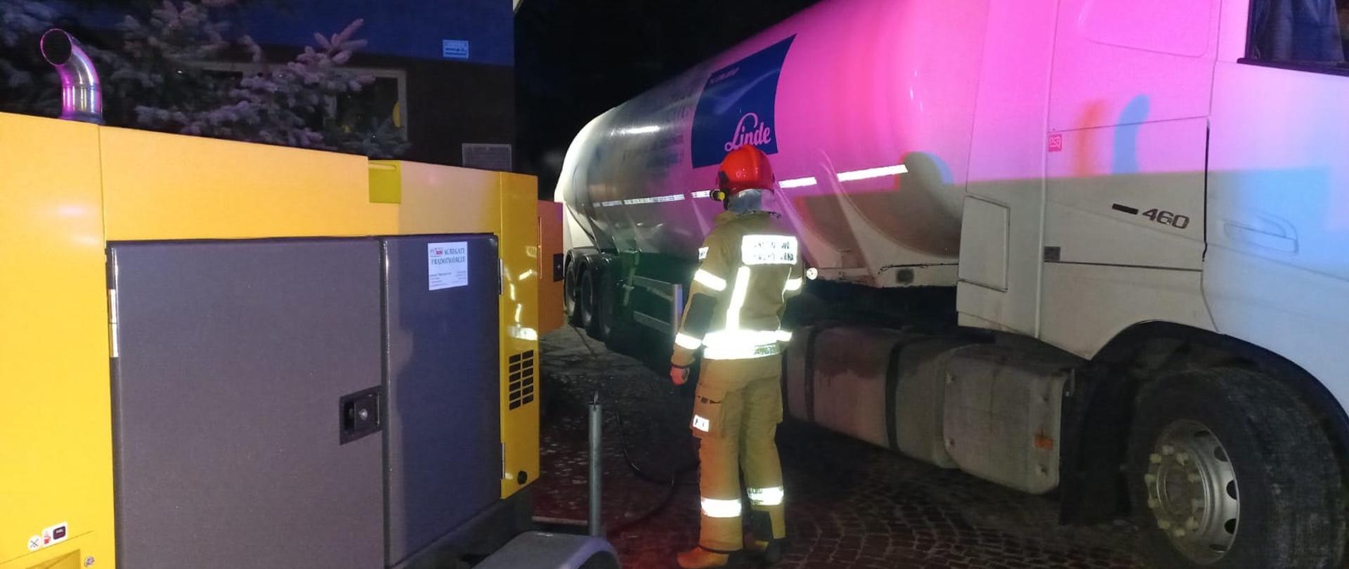 Zdjęcie przedstawia strażaków pomagających w uzupełnieniu tlenu medycznego dla szpitala.
W tle budynek i drzewa.
