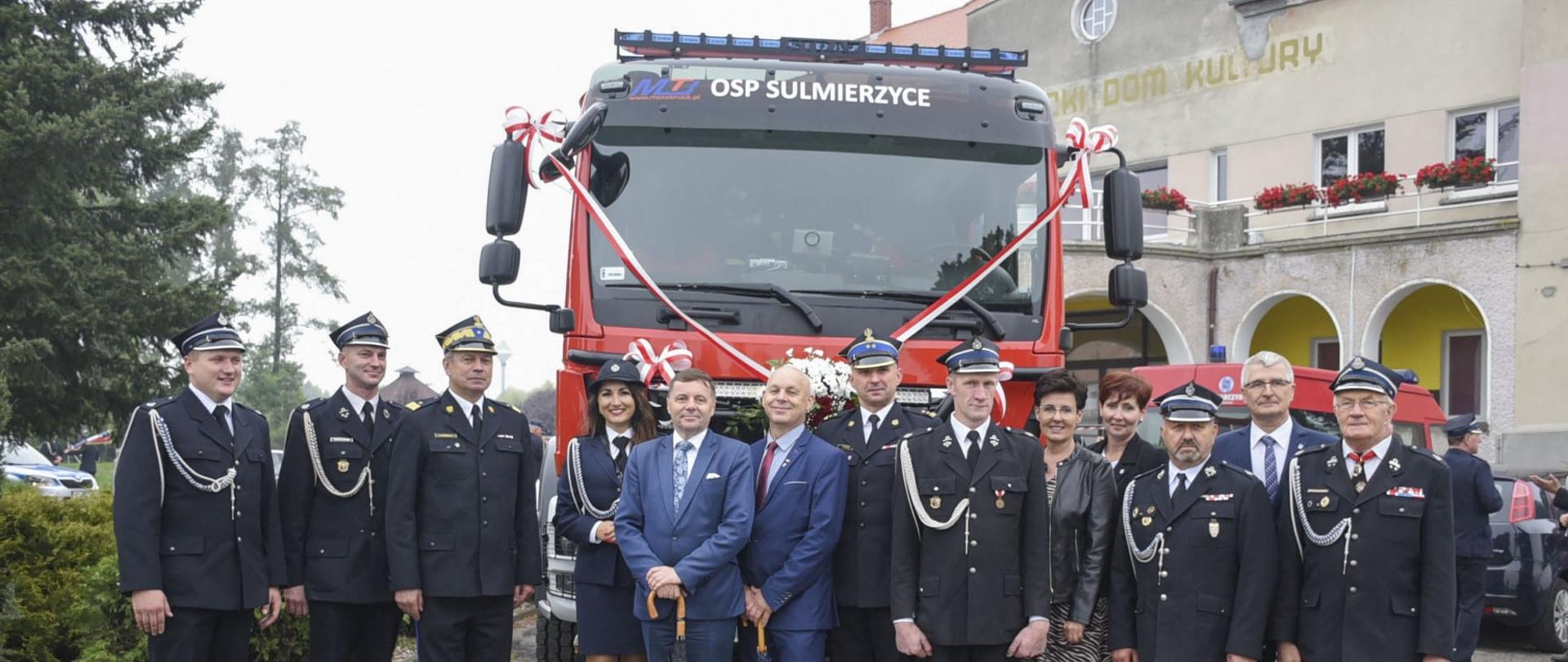Grupowe zdjęcie strażaków PSP, druhów OSP i osób cywilnych na tle samochodu ratowniczo - gaśniczego OSP, w tle budynek Domu Kultury