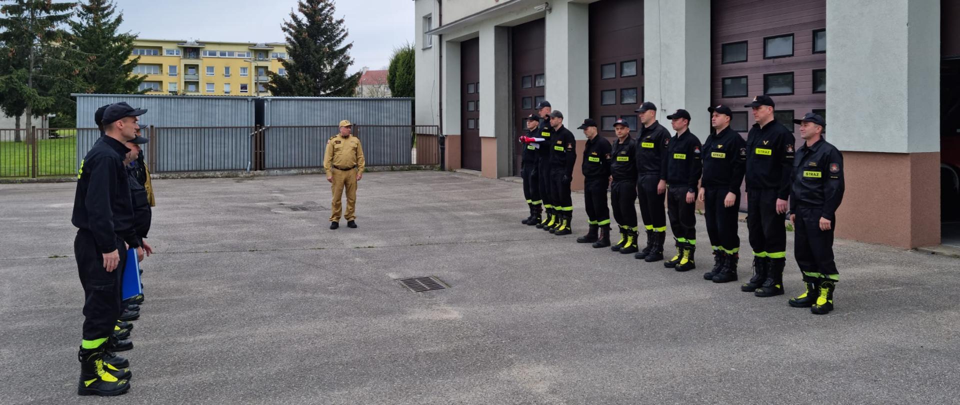 Fotografia przedstawia uroczyste przyjęcie służby z okazji Dnia Flagi Rzeczypospolitej Polskiej. Strażacy z dwóch zmian służbowych stoją na przeciw siebie z prawej strony stoi poczet flagowy składający się z trzech strażaków, z których jeden trzyma złożoną Flagę Państwową w kolorze biało czerwonym.