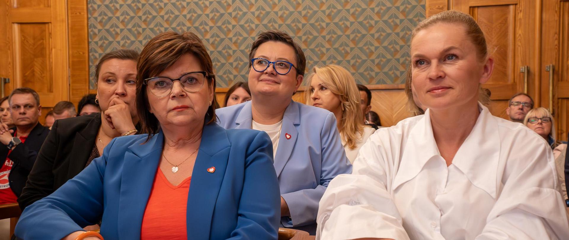 Widok z przodu na dużą salę, na ustawionych w rzędy krzesłach siedzą ludzie. W pierwszym rzędzie minister Barbara Nowacka i minister Izabela Leszczyna. Za nimi wiceminister Katarzyna Lubnauer i Paulina Piechna-Więckiewicz