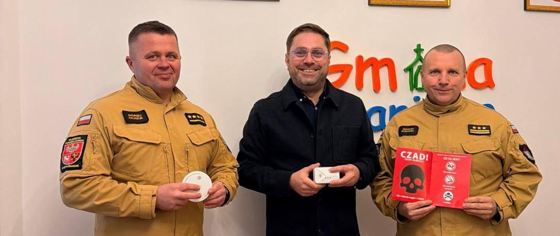 Na zdjęciu w gabinecie trzy osoby. Dwóch strażaków w jasnych mundurach, w środku wójt gminy Braniewo, trzymają w rekach czujki tlenku węgla. W tle na ścianie herb powiatu braniewskiego oraz godło Polski.