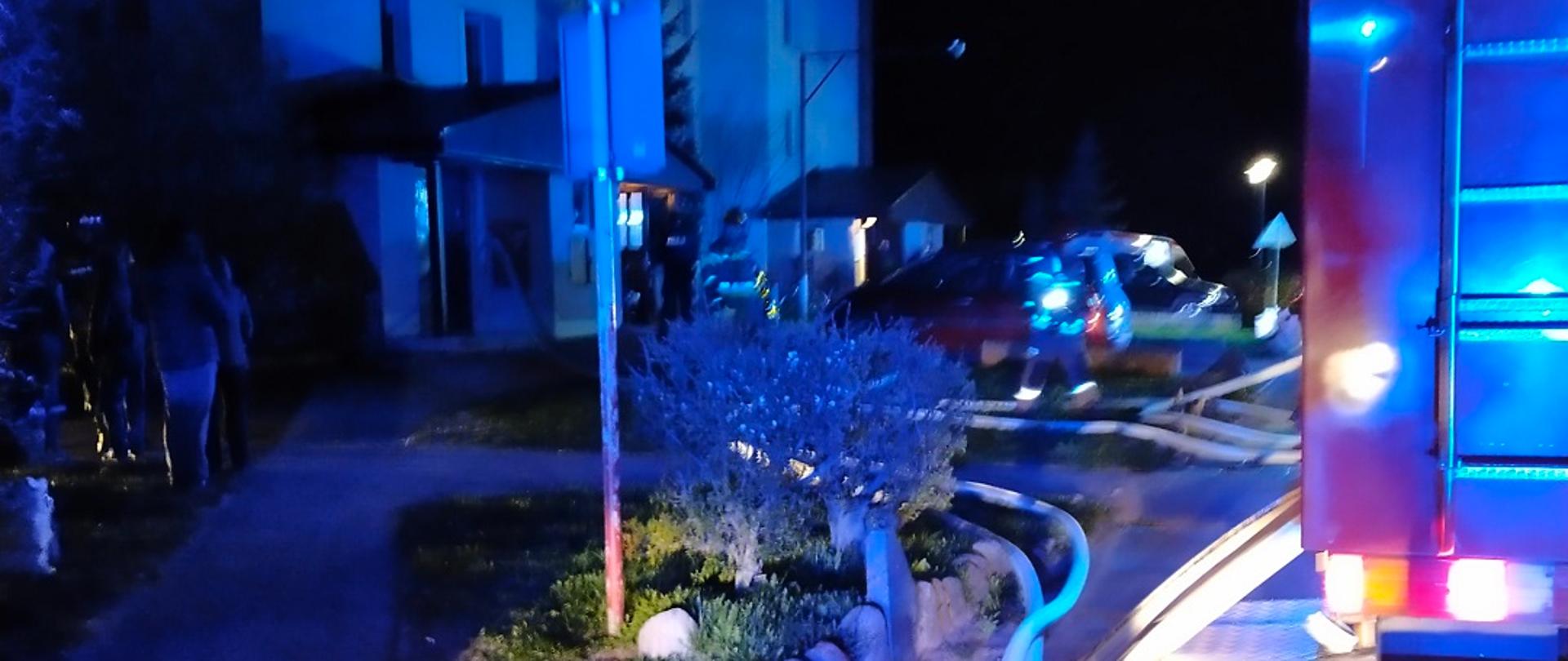 Zdjęcie obrazuje cześć samochodu pożarniczego węże strażackie część bloku strażaków i osoby cywilne w porze nocnej