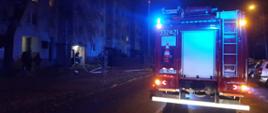 Zdjęcie wykonane w porze nocnej. Na zdjęciu widać ulicę. Na niej ustawiony jest pojazdy ratowniczo - gaśniczy. Pojazd w kolorze czerwonym ze srebrnymi elementami zabudowy. Samochód dodatkowo włączony ma niebieskie alarmowe sygnały świetlne. Obok pojazdu w pewnej odległości po lewej stronie widać blok mieszkalny czteropiętrowy. W jednym z jego mieszkań wybuchł pożar. Od pojazdu do wejścia do klatki schodowej rozłożona jest linia gaśnicza z węży pożarniczych. Przed blokiem dodatkowo widać stojących strażaków. 
