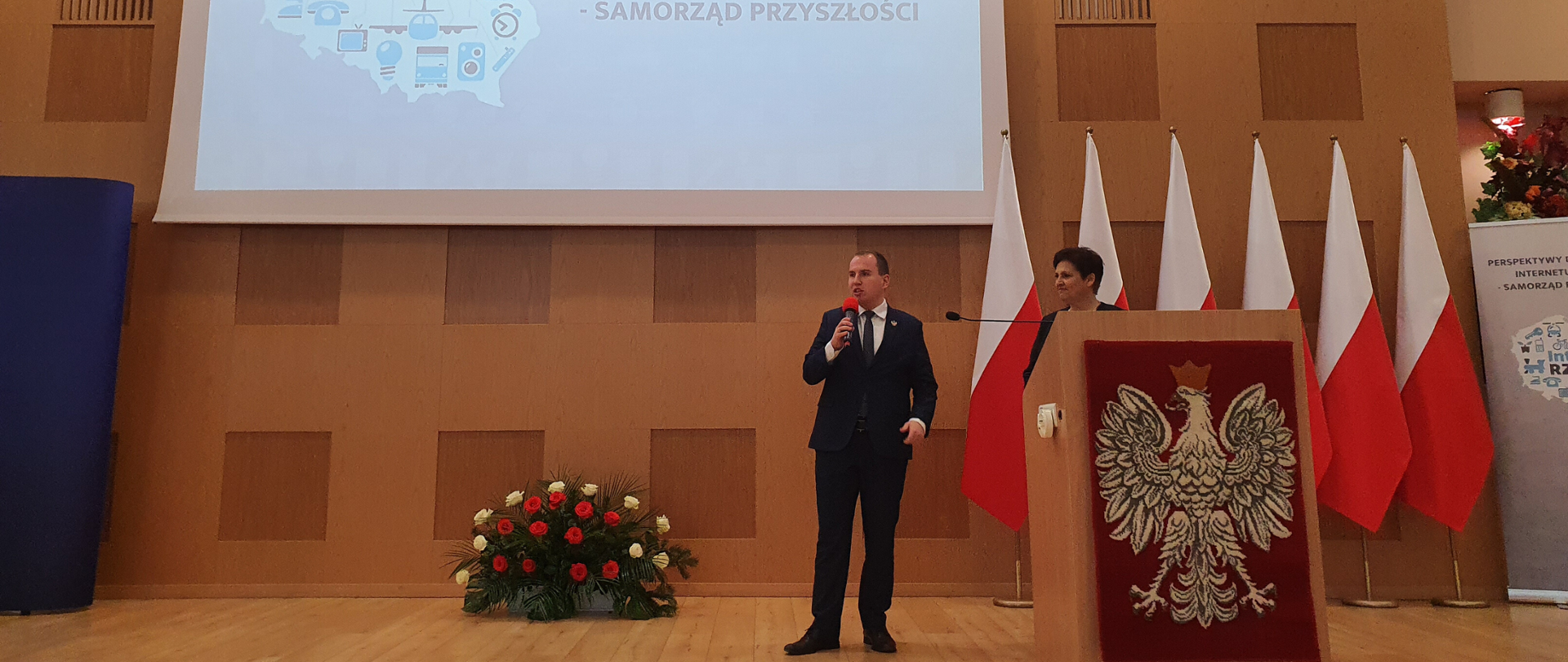 Wiceminister cyfryzacji stoi z mikrofonem o obok mównicy. Obok niego stoi Halina Szymańska, szef Kancelarii Prezydenta RP. W tle polskie flagi. Nad nimi wisi ekran z nazwą konferencji i jej logotypem.