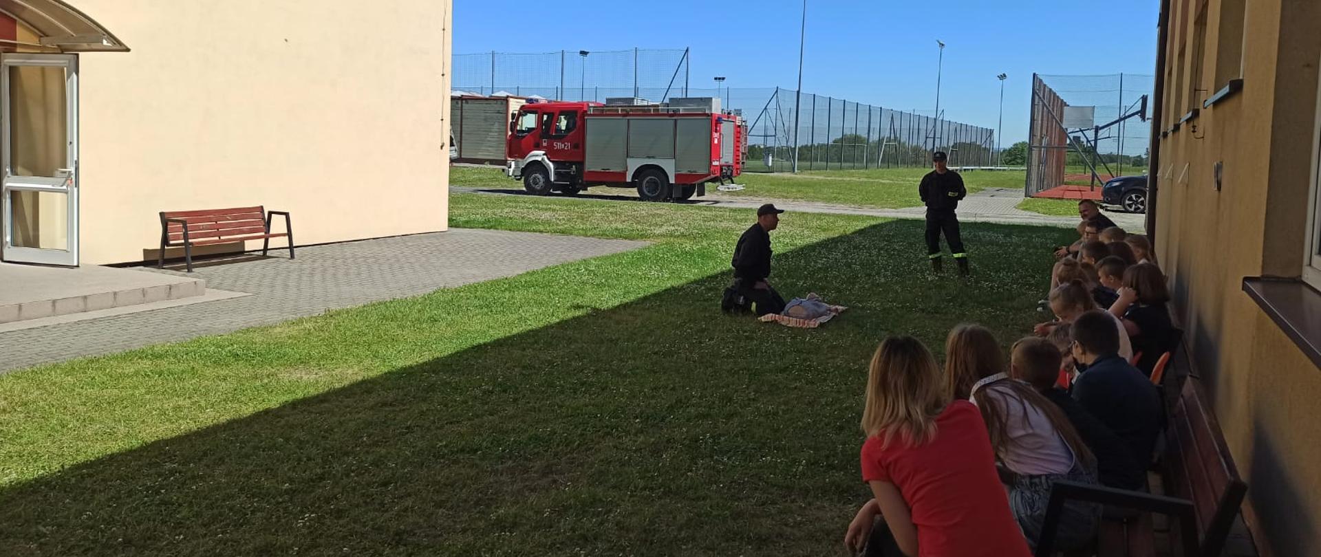 Zdjęcie wykonane jest przed budynkiem szkoły. Pod ścianą w cieniu znajdują się siedzące na krzesłach dzieci, które przyglądają się dwóm strażakom. Strażacy wykonują pokazy udzielania kwalifikowanej pierwszej pomocy na manekinie. W tle zdjęcia znajduje się wóz strażacki.