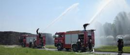 Na zdjęciu widać strażaków, którzy z dwóch samochodów pożarniczych podają wodę z działka. W tle widoczny jest skład palet i torowisko.
