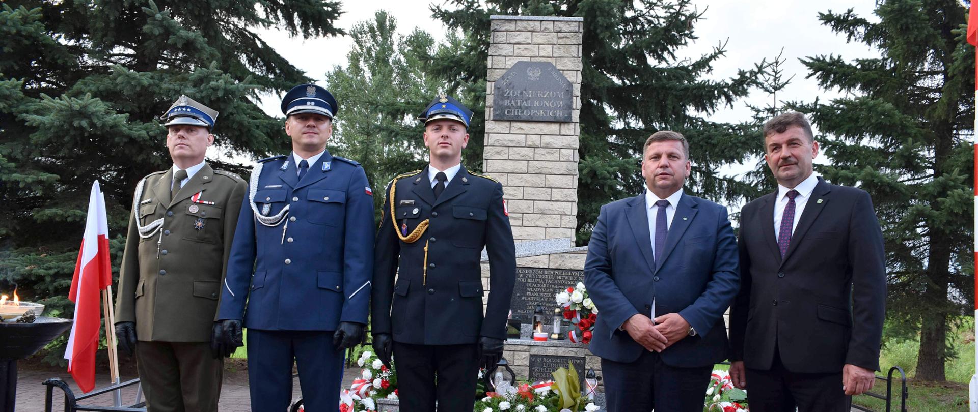 Zdjęcie przedstawicieli służb mundurowych oraz władz samorządowych stojących przed pomnikiem.
Od lewej strony stoi trzech mężczyzn w mundurach: żołnierz w zielonym mundurze, funkcjonariusz w niebieskim mundurze oraz funkcjonariusz w czarnym mundurze galowym. Obok nich, po prawej stronie, stoją dwaj mężczyźni w eleganckich garniturach z krawatami W tle znajduje się kamienny pomnik z tablicą upamiętniającą, otoczony zielonymi drzewami oraz wieńcami kwiatów u podstawy.