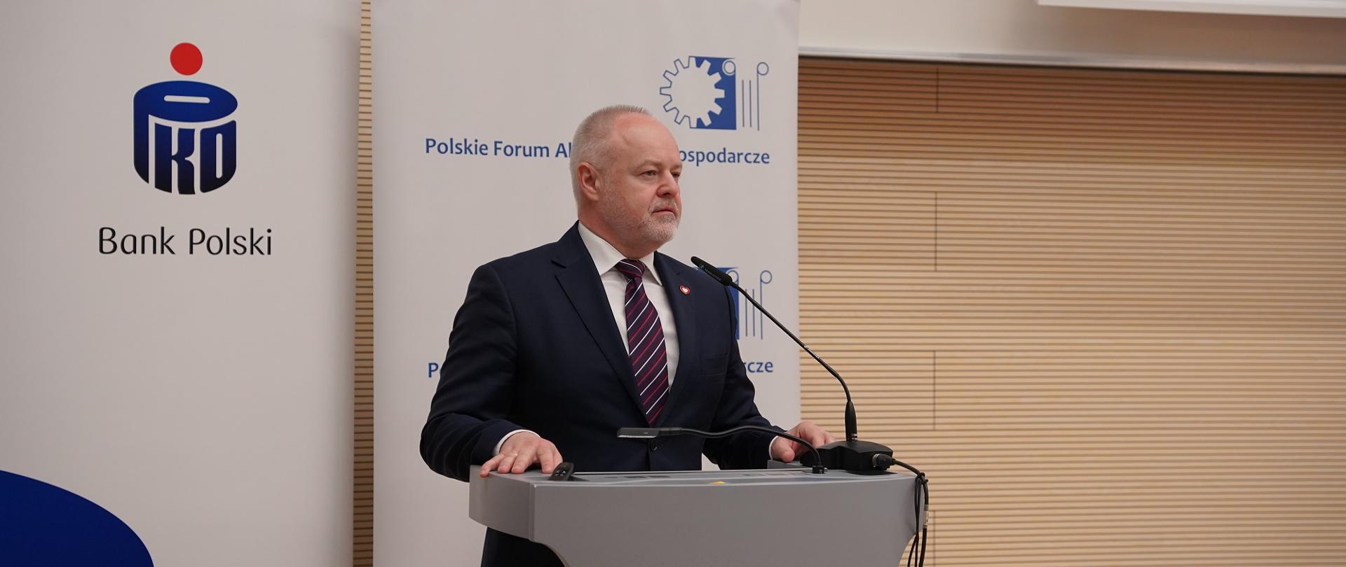Wiceminister Gzik stoi za szarą mównicą, za nim pod ścianą z poziomych drewnianych listewek banery z napisem PKO Bank Polski i Polskie Forum Akademicko-Gospodarcze.