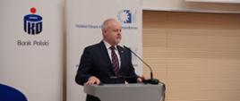 Wiceminister Gzik stoi za szarą mównicą, za nim pod ścianą z poziomych drewnianych listewek banery z napisem PKO Bank Polski i Polskie Forum Akademicko-Gospodarcze.