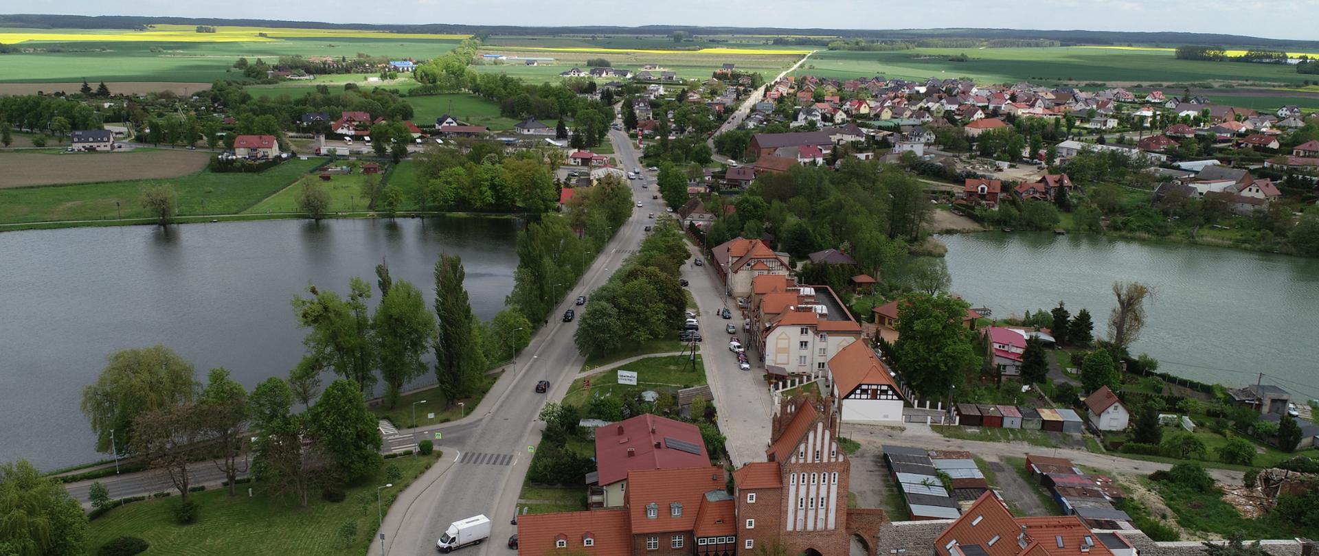 Zdjęcie wykonano z powietrza. Na zdjęciu widać panoramę miasta, przez środek biegnie główna droga po obu stronach drogi są zbiorniki wodne (jeziorka) i typową zabudowę miejską średniego miasta z przeważającymi domami jedno lub kilkurodzinnymi. W oddali rozciąga się widok na zielone i żółte połacie pól.