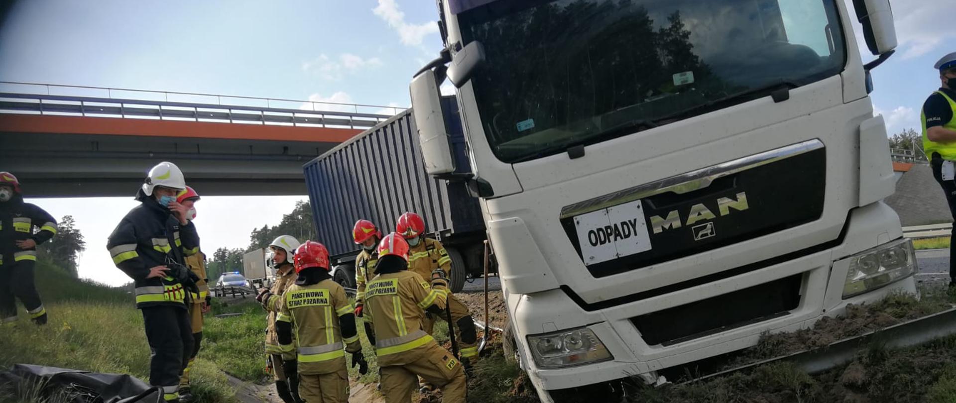 Zdjęcie przedstawia akcję ratowniczą na S8. Na zdjęciu widać pojazd ciężarowy, który częściowo wypadł z jezdni. Po lewej widzimy strażaków biorących udział w akcji. Zdjęcie wykonane w porze dziennej na poboczu drogi S8.