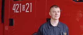 Zdjęcie przedstawia mł. asp. Krystiana Drzewickiego stojącego przy samochodzie pożarniczym w pomieszczeniach garażowych Jednostki Ratowniczo – Gaśniczej w Człuchowie