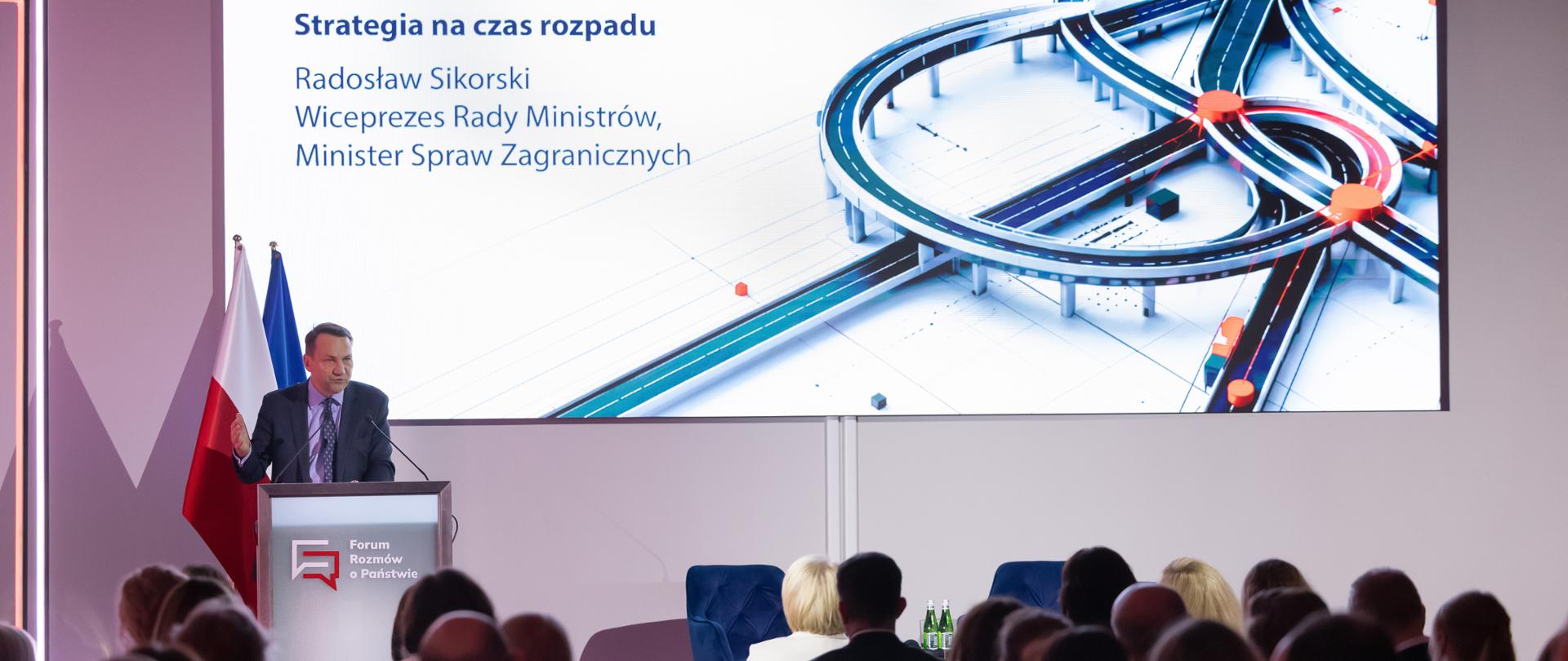 Wicepremier, minister spraw zagranicznych wygłasza przemówienie w Krajowej Szkole Administracji Publicznej stojąc za mównicą z napisem "Forum Rozmów o Państwie" na tle flagi Polski i Unii Europejskiej. Za ministrem prezentacja ze słowami "Sprawcze Państwo. Strategia na czas rozpadu" 