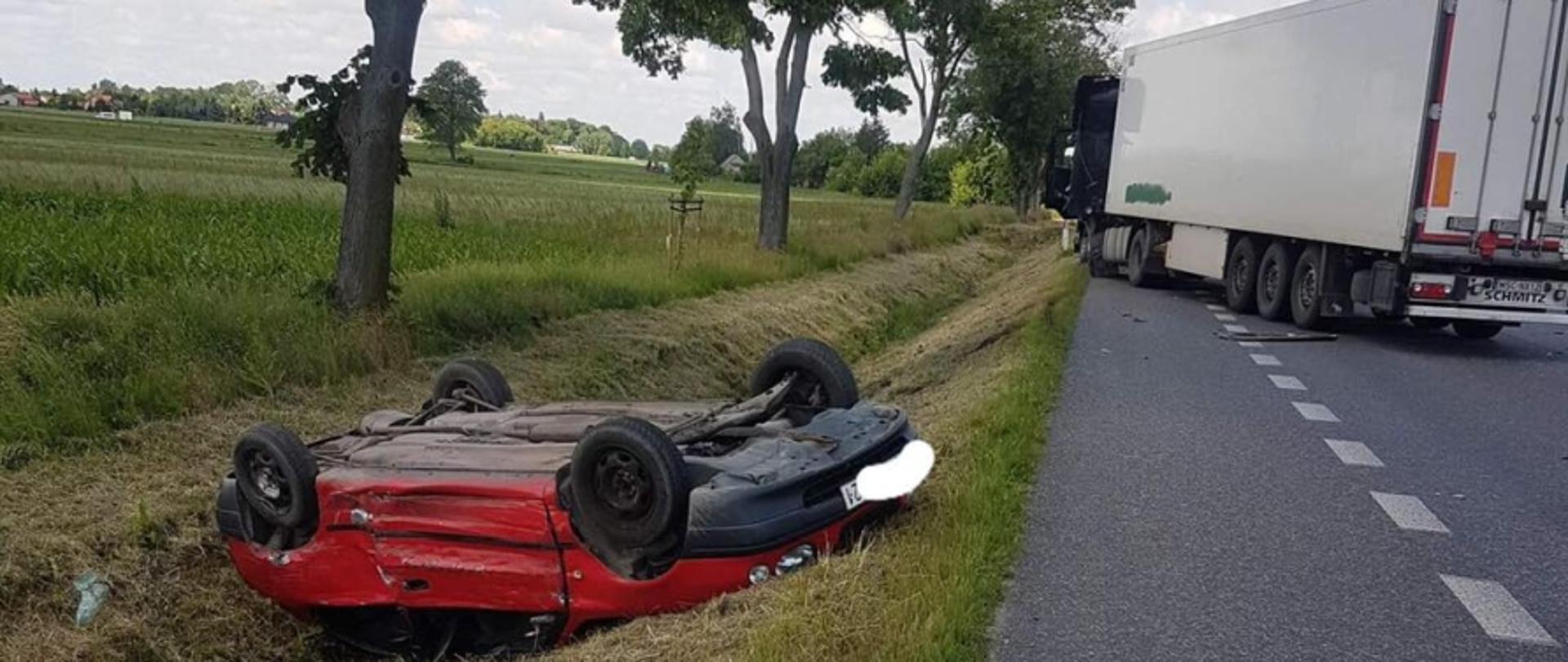 Na zdjęciu na pierwszym planie widoczny samochód osobowy koloru czerwonego leżący na dachu w przydrożnym rowie. Na drugim planie zestaw - ciągnik siodłowy z naczepą.