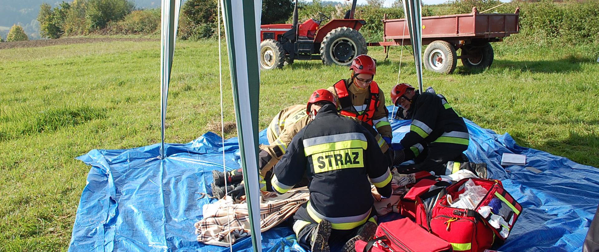 Zdjęcie przedstawia strażaków w umundurowaniu specjalnym wykonujących założenie z ratownictwa medycznego. Działania prowadzone są na skraju lasu. W tle widoczny jest ciągnik rolniczy z przyczepą jako element pozoracji.