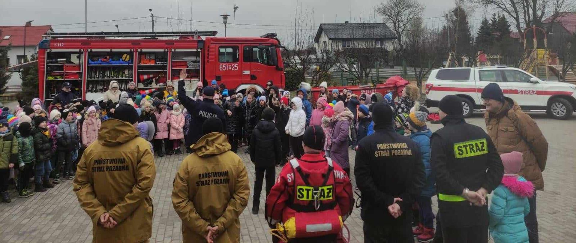 Zdjęcie przedstawia strażaków oraz policjanta stojących przed dziećmi obok wozów strażackich