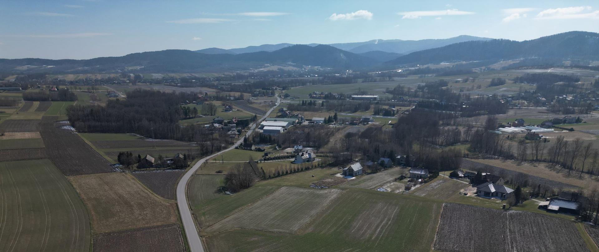 Węzeł Inwałd, teren budowy przyszłego węzła Inwałd, w dolnej części zdjęcia widoczne przeważnie pola, dalej zabudowania, w oddali Beskidy. Przez znaczną długość zdjęcia prowadzi lokalna droga