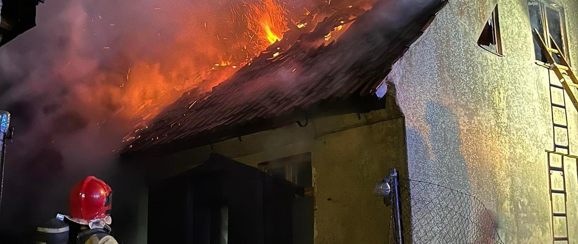 Na zdjęciu widać strażaka podającego wodę na płonący dach budynku