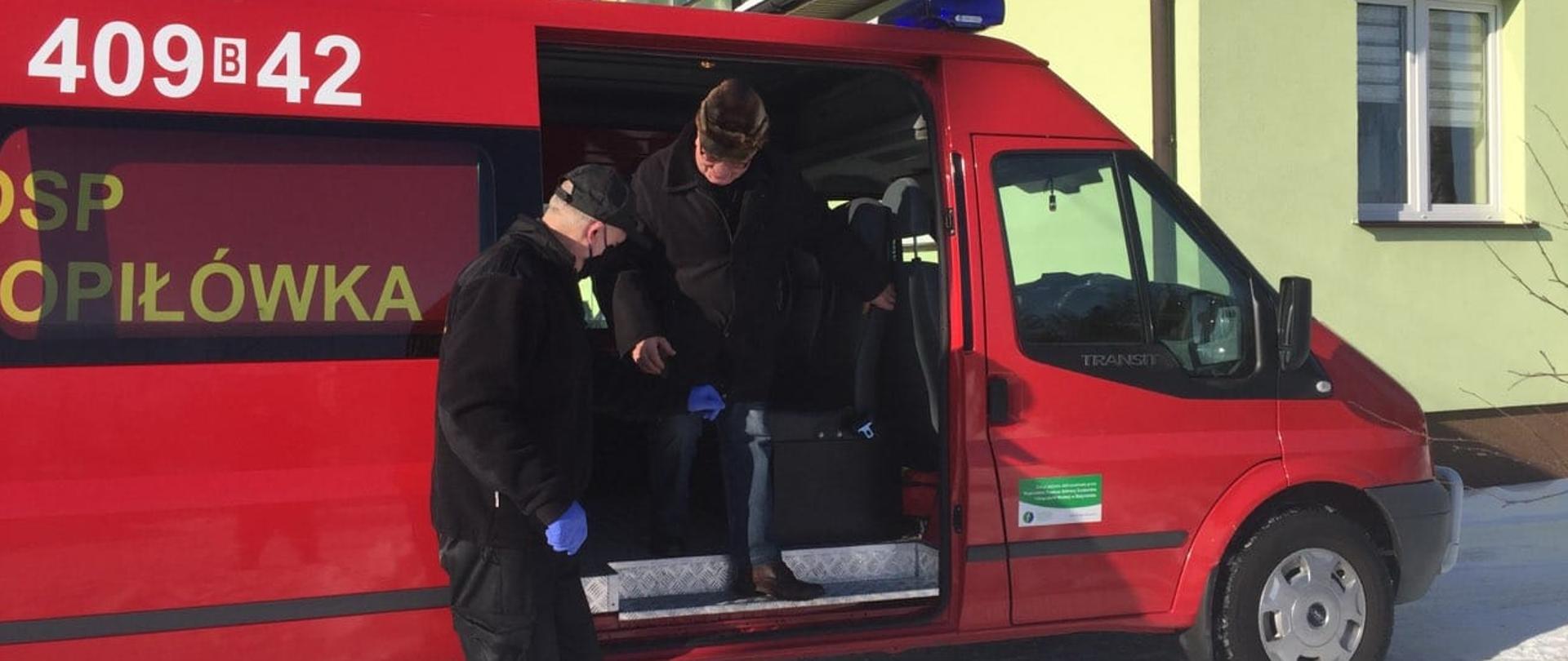 
W tle widać samochód pożarniczy podczas dowozu osoby do punktu szczepień oraz strażaka pomagającemu starszej osobie w wyjściu z pojazdu.
