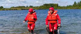 Scena na jeziorze przedstawiająca dwóch ratowników, stojących w płytkiej wodzie i wychodzących w kierunku brzegu. Obaj ubrani są w czerwone kombinezony i kaski oraz pomarańczowe kamizelki ratunkowe. Ratownik po lewej trzyma w dłoni linę. Ratownik po prawej, uśmiechnięty, niesie w prawej ręce parę płetw. Za nimi rozpościera się jezioro, a w oddali widoczny jest brzeg porośnięty rzadkimi krzakami i drzewami. Nad nimi błękitne niebo z białymi chmurami.