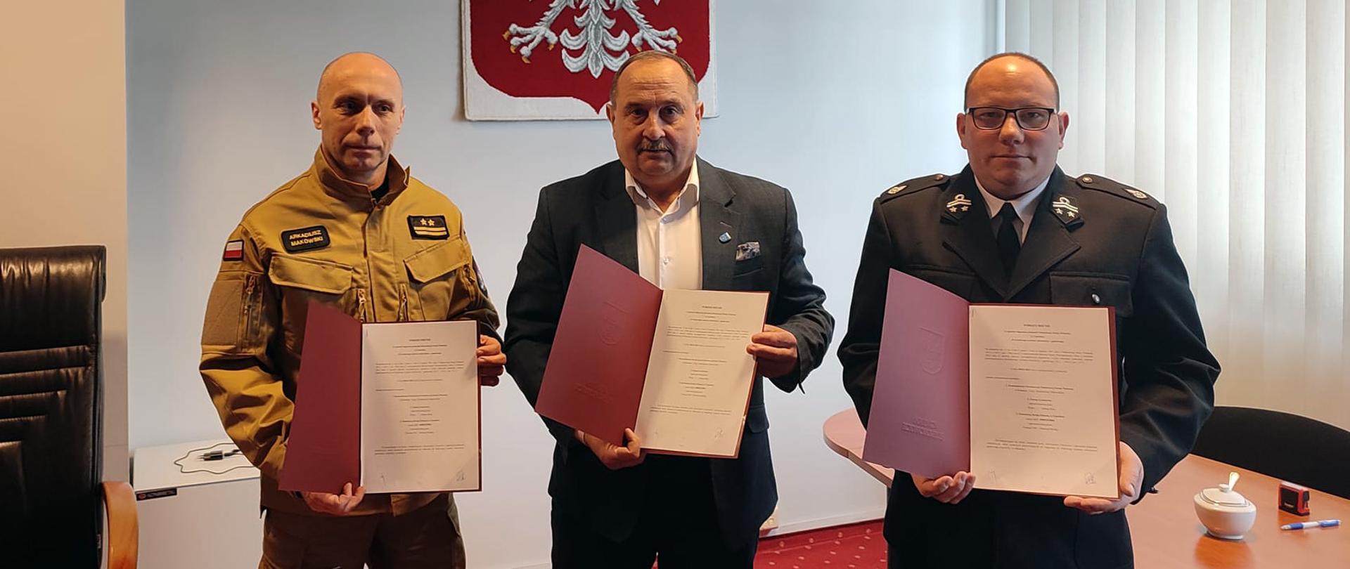 trzy osoby trzymają w ręku dokumenty w czerwonej teczce jedna osoba ubrana jest w mundur państwowej straży pożarnej druga w mundur galowy bez sznura ochotniczych straży pożarnych i trzecia w garniturze bez krawata w tle na ścianie duże godło polski a po jego lewej stronie herb gminy Łyszkowice pod herbem skórzany fotel po prawej stronie okno przysłonięte pionowymi żaluzjami przy oknie stół
