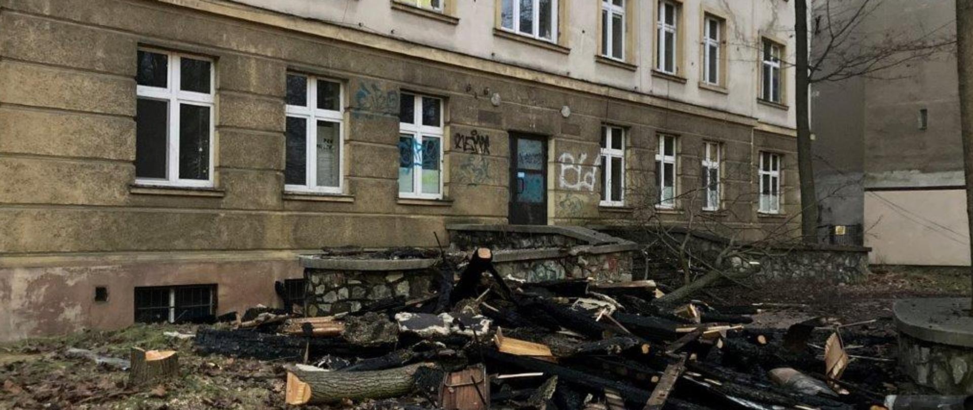 Zdjęcie przedstawia budynek opuszczonego szpitala gdzie doszło do pożaru. Na placu widać pryzmę spalonych elementów drewnianych konstrukcji dachowej.