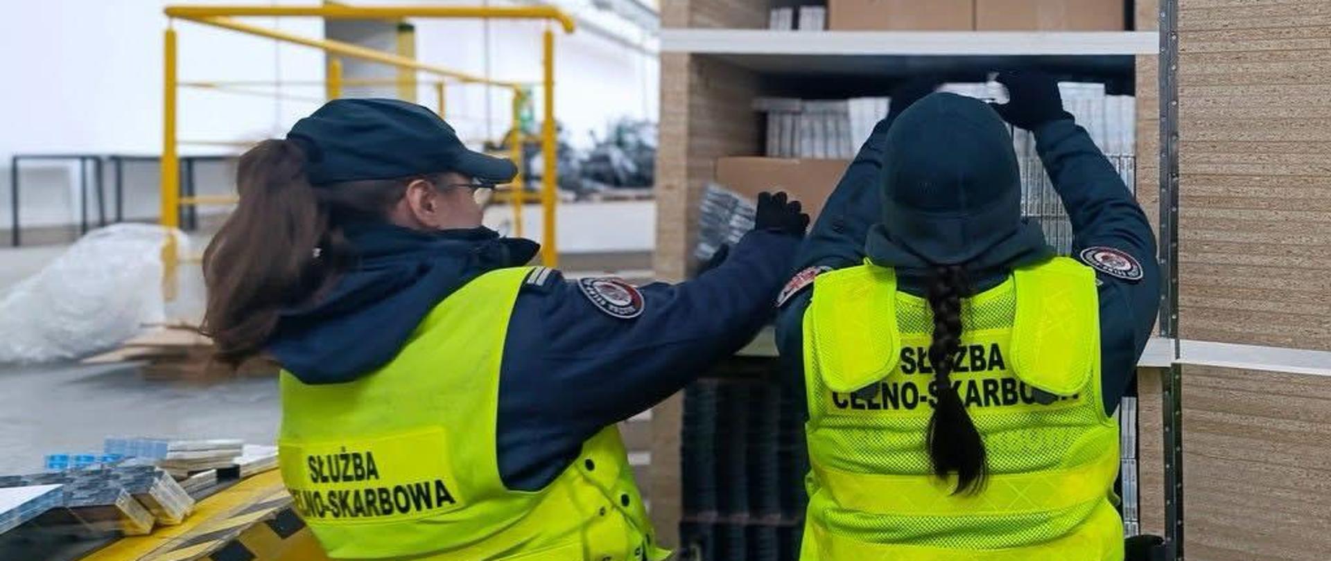 Dwie osoby w kamizelkach z napisem „Służba Celno‑Skarbowa” stoją przy metalowym regale i sprawdzają ułożone na półkach opakowania; w tle widać elementy hali magazynowej oraz żółtą barierkę.