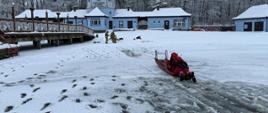 Strażacy w czerwonych ubraniach wypornościowych do pracy w wodzie ćwiczą podejmowanie osoby poszkodowanej, pod którą załamał się lód i wpadła do wody. Na zdjęciu strażacy na saniach lodowych. Zamarznięte jezioro w zimowej aurze. Strażacy wykonują manewr sciągania sań lodowych na brzeg jeziora, poszkodowany jest osobą przytomną.