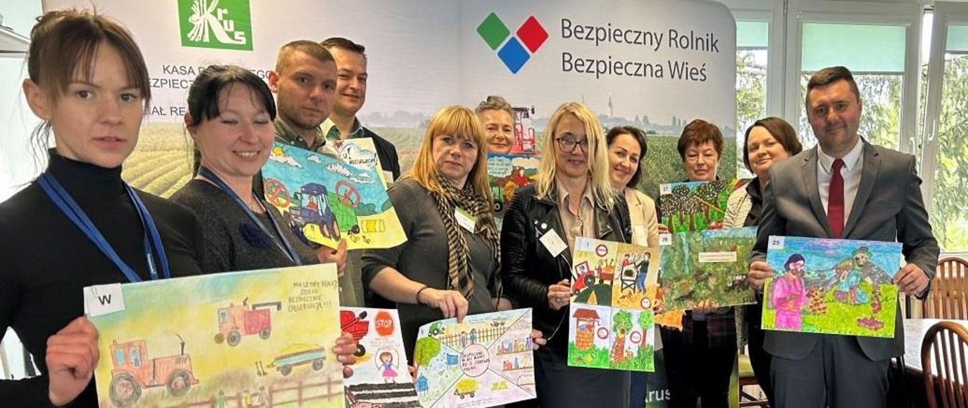 Grupa osób pozuje do zdjęcia z pracami plastycznymi w rękach na tle baneru KRUS
