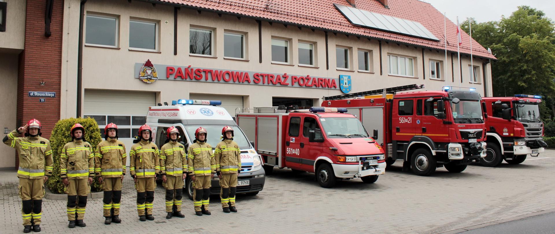 Strażacy w ubraniach specjalnych stoją w szyku przed wozami gaśniczymi jeden z nich salutuje