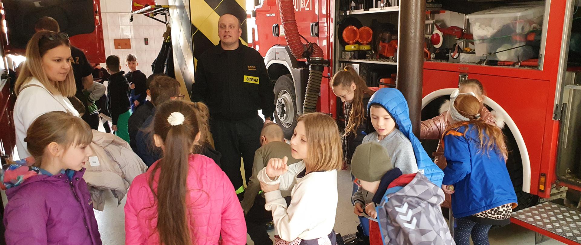Na zdjęciu widać dzieci ze szkoły podstawowej które znajdują się na garażu jednostki straży pożarnej. Zwrócone są One plecami do zdjęcia a przed nimi stoi strażak - dowódca zmiany w ubraniu koszarowym który omawia sprzęt. Po lewej stronie znajduje się Pani nauczycielka, a po prawej samochód strażacki z otwartymi skrytkami. 