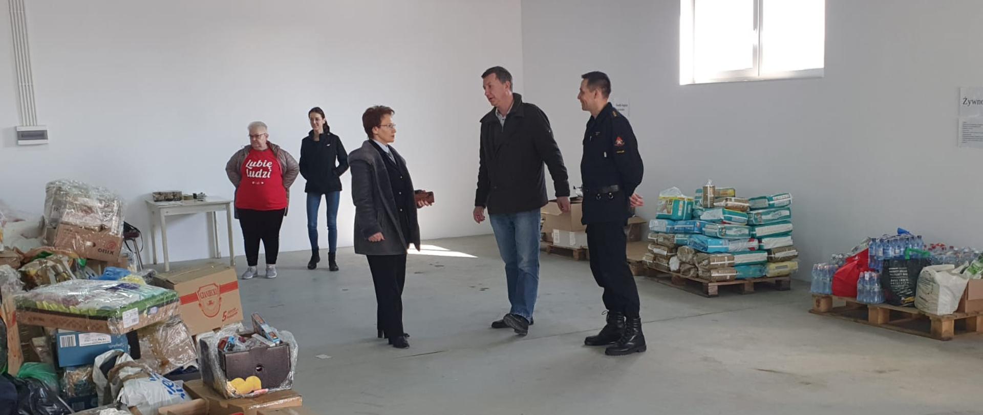 Na zdjęciu w powiatowym magazynie pomocy wewnątrz hali pomiędzy składowanymi na paletach darami dla obywateli Ukrainy widoczni starosta gorlicki i zastępca komendanta powiatowego PSP w Gorlicach oraz trzy osoby cywilne.