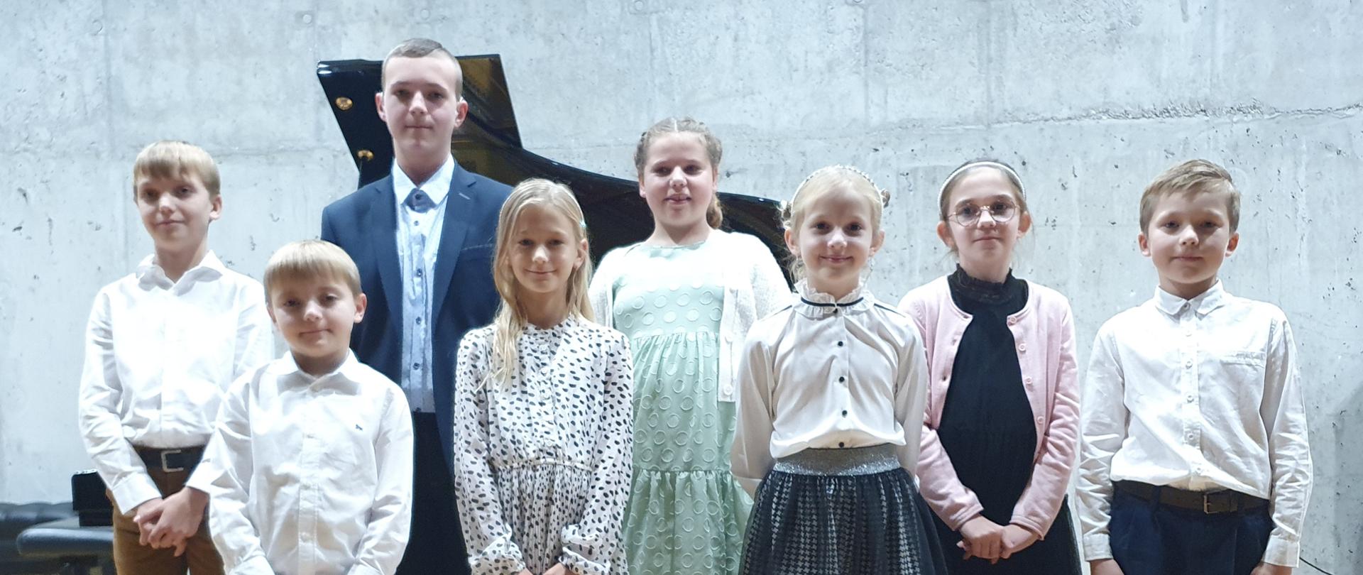 Grupa uczniów stoi na scenie, fortepian w tle