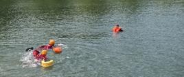 Dwóch strażaków w specjalistycznych ubraniach płynących do osoby poszkodowanej, która jest w kamizelce ratunkowej na środku zbiornika wodnego