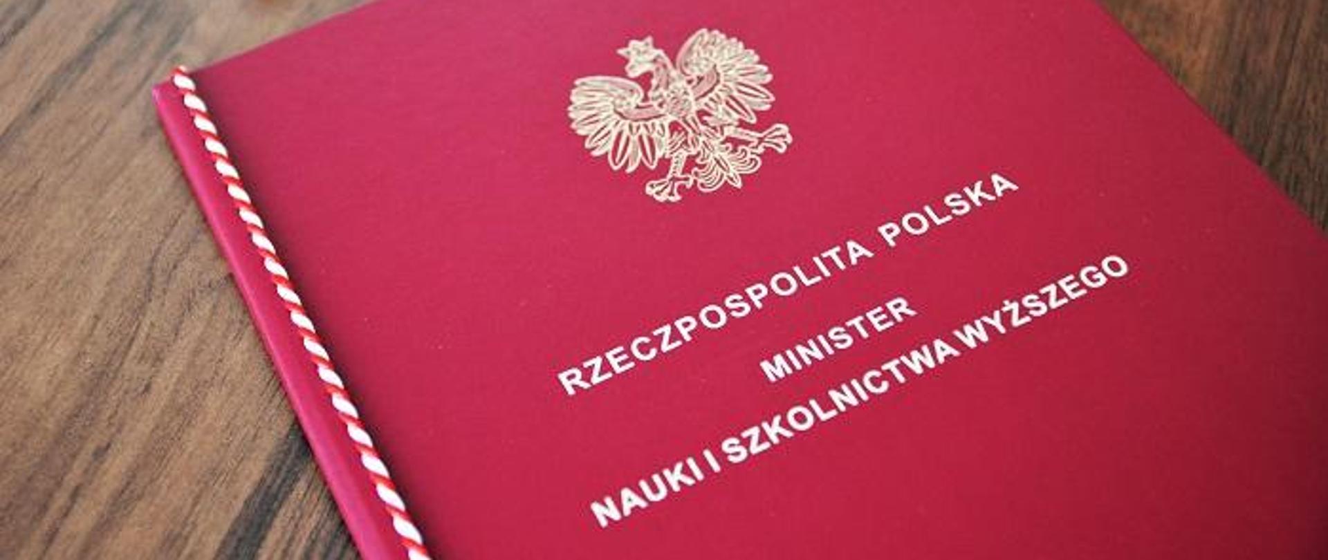 Na drewnianym blacie lezy czerwona teczka z napisem Rzeczpospolita polska - minister nauki i szkolnictwa wyższego