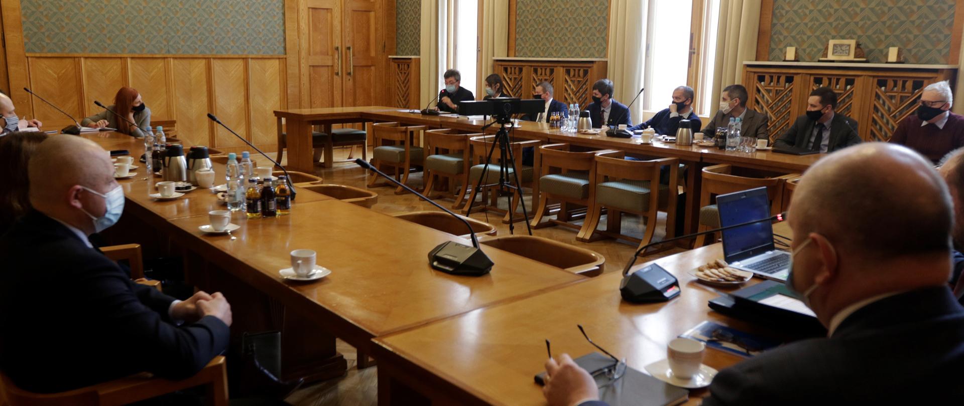 Na zdjęciu grupa osób osób, głównie mężczyzn siedzących przy stole ustawionym w podkowę. Mężczyźni są ubrani w garnitury. Na twarzach mają maseczki. Na stole stoją mikrofony, napoje i filiżanki., 