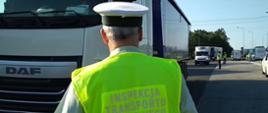 Na pierwszym planie umundurowany inspektor ITD stoi przed kabiną ciężarówki zatrzymanej do kontroli. W tle widać pozostałych inspektorów, którzy prowadzą kontrole trzeźwości kierujących.