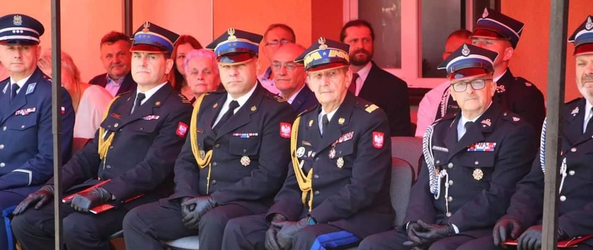 Kolorowa fotografia wykonana w pogodny dzień na zewnątrz. Przedstawia strażaków PSP i OSP oraz osoby cywilne podczas uroczystości 140 lecia OSP Jasionów. Strażacy występują w umundurowaniu wyjściowym i siedzą