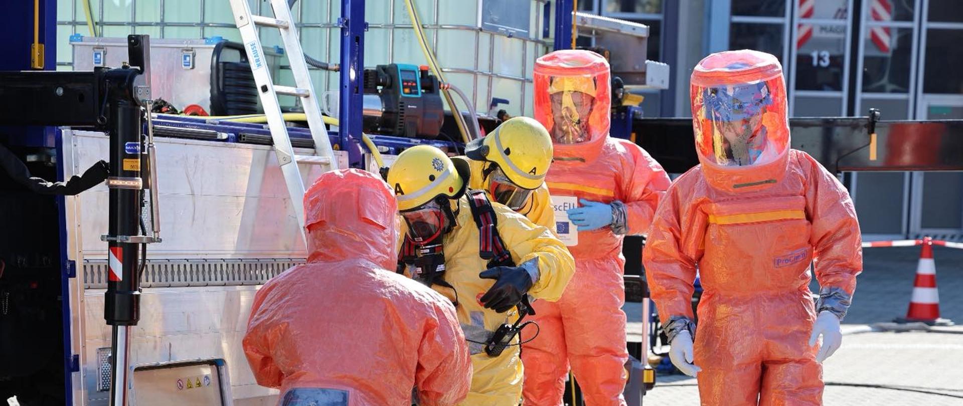 Na zdjęciu widać grupę pięciu osób ubranych w specjalne, szczelne kombinezony ochronne. Trzy osoby mają pomarańczowe kombinezony z przezroczystymi osłonami na twarzy i zielonymi butami, a dwie osoby są ubrane w żółte kombinezony z żółtymi hełmami i maskami. Wszyscy biorą udział w pozorowanej akcji związanej z zagrożeniem chemicznym i biologicznym. Stoją obok metalowego kontenera z różnym sprzętem technicznym i wężami. W tle widać budynki i pomarańczowe pachołki bezpieczeństwa. Scena odbywa się na zewnątrz, na brukowanym placu, w słoneczny dzień.