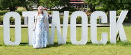 Paulina Nadworska, młoda dziewczyna w niebiesko-białej sukience w kwiaty stoi w plenerze przed dużymi literami „Otwock" ustawionymi na trawie.