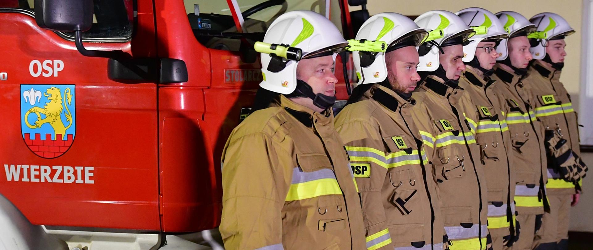 sześciu strażaków w ubraniach specjalnych stojących przed samochodem gaśniczym, obok drzwi kierowcy samochodu na których widnieje napis OSP Wierzbie, 
