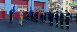 Druhów OSP stojących w szeregu. Niektórzy z nich trzymają sztandar. Naprzeciwko ich stoi strażak PSP który omawia ceremoniał pożarniczy. Obok niego stoi drugi strażak ze sztandarem w rękach.