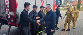 Wiceminister MSWiA oraz Zastępca Komendanta Głównego PSP i Komendant Powiatowy PSP w Wołominie wita się ze strażakami stojącymi w szeregu przy samochodzie strażackim.