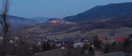Zdjęcie zrobione z wzniesienia zlokalizowanego naprzeciw góry na której zboczu widoczne są płomienie palącej się trawy. Poniżej pożaru w dolinie widoczne są zabudowania miejscowości.