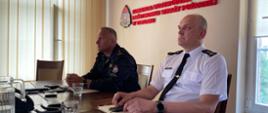 Na zdjęciu dwóch zastępców komendanta wojewódzkiego w trakcie słuchania wykładu on-line. Siedzą na sali konferencyjnej, w umundurowaniach - jeden letnim wyjściowym, drugi w dowódczo-sztabowym. Za nimi na ścianie logi i napis firmowy.