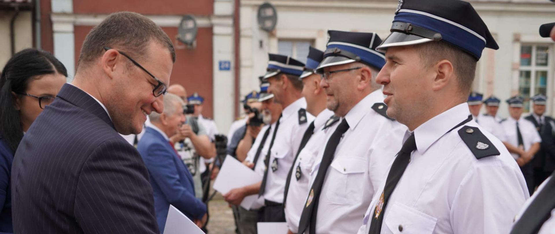 Zdjęcie przedstawia wręczenie dofinansowania. Mężczyzna w garniturze wręcza druhowi OSP dokument i podają sobie ręce.