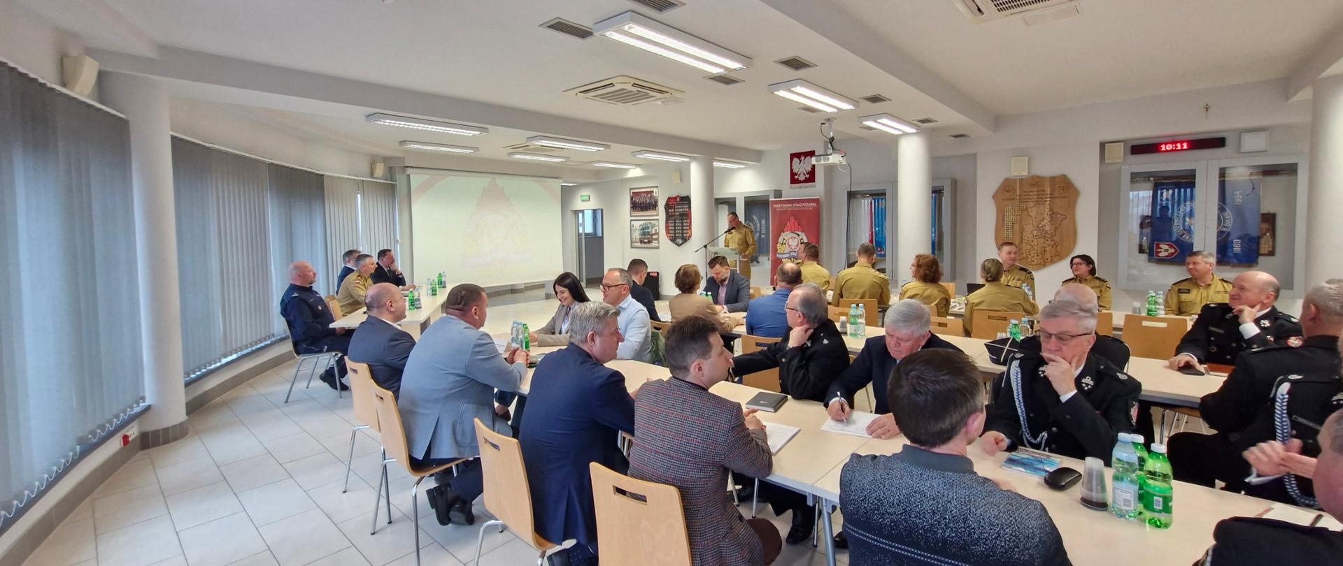 Zdjęcie przedstawia strażaków oraz zaproszonych gości na naradzie.. Jeden strażak prezentuje dokonania KP PSP w Bochni w 2024 r. 